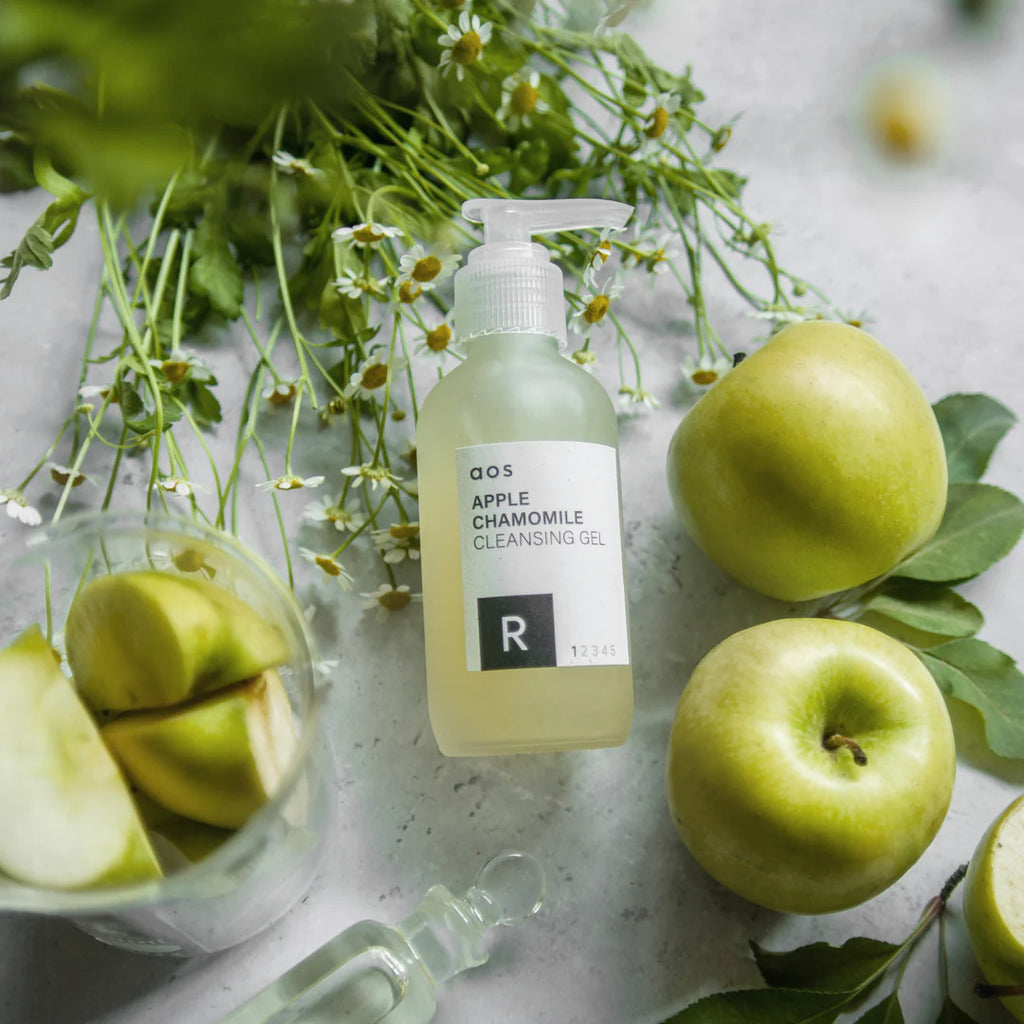 Bottle of chamomile apple cleansing gel surrounded by fresh apples and greenery on a marble surface.