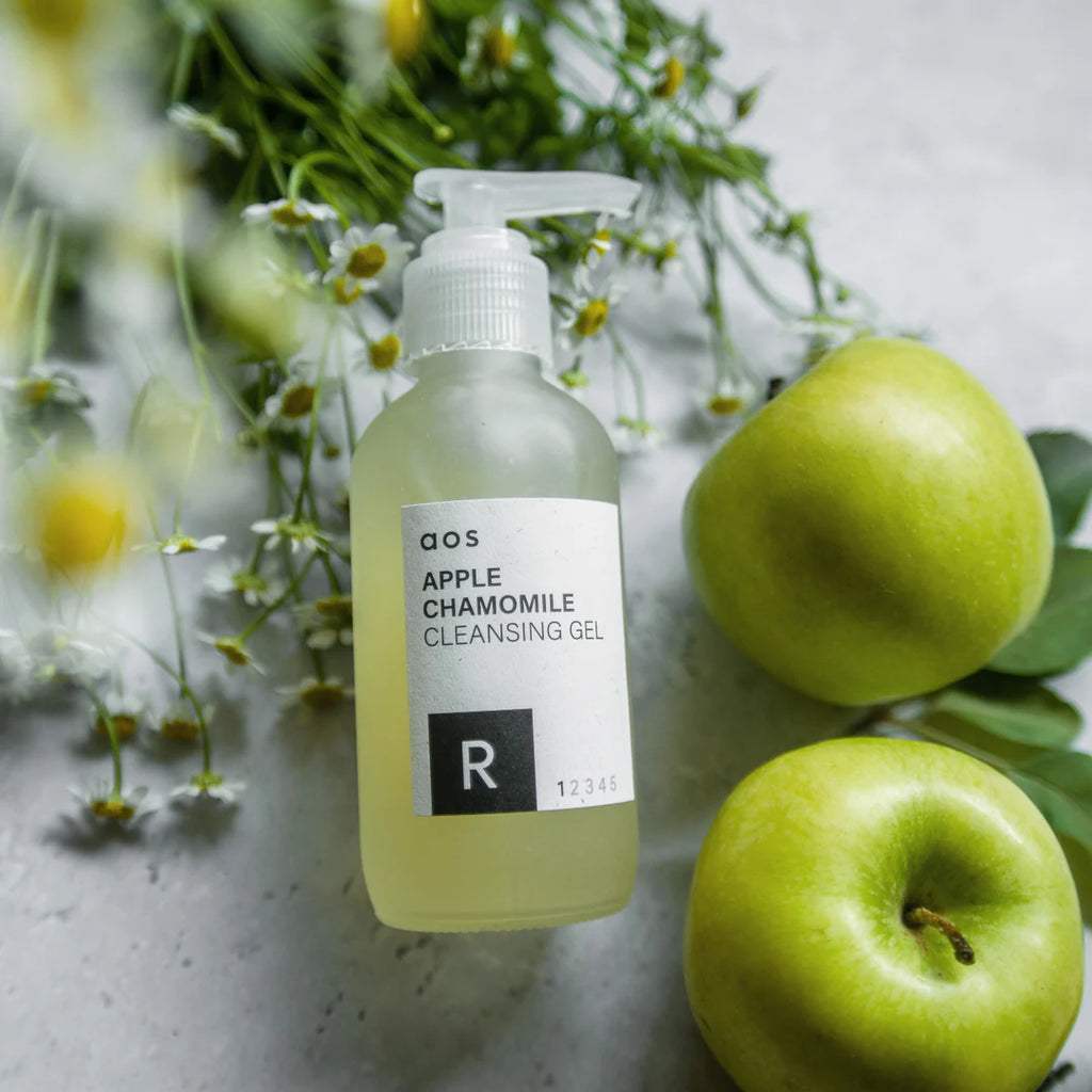 Bottle of apple chamomile cleansing gel with fresh green apples and chamomile flowers on a marble surface.
