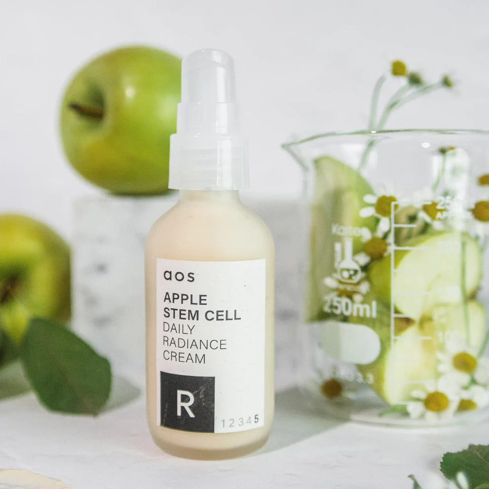 A bottle of apple stem cell radiance serum with fresh apples and a beaker of plants in the background.