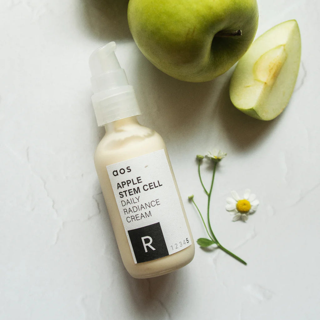 Bottle of apple stem cell daily radiance cream placed beside a green apple and a small white flower on a marble surface.