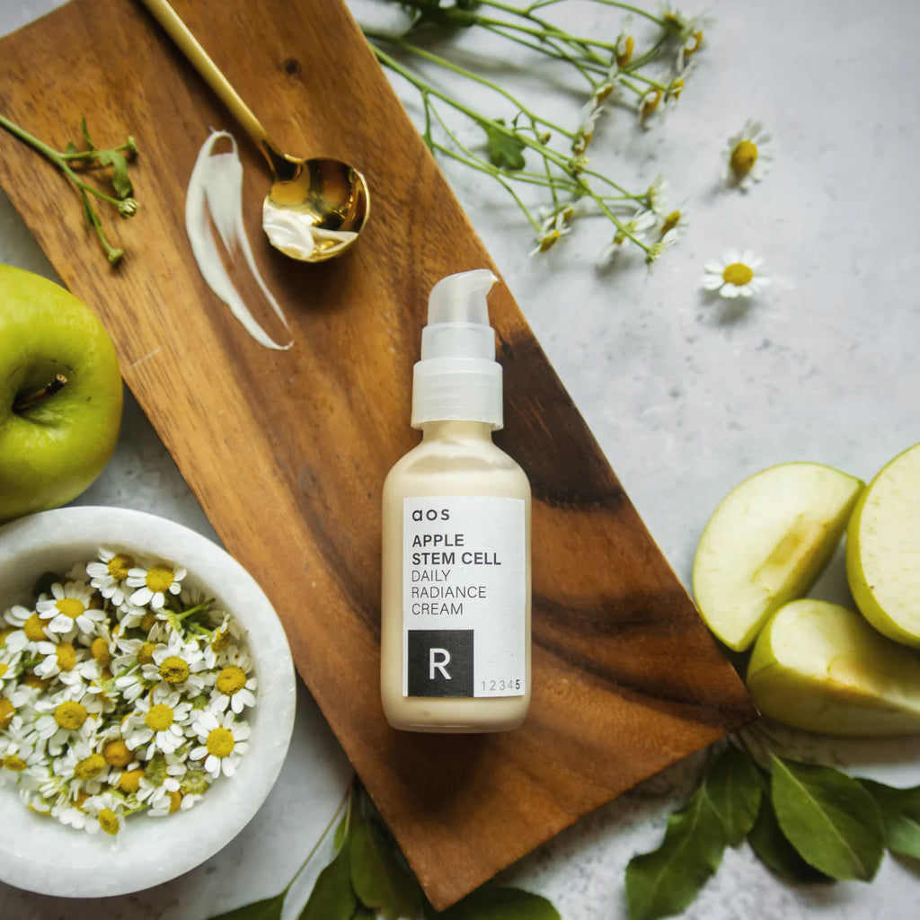 A bottle of apple stem cell radiance cream on a wooden board surrounded by fresh apples, chamomile flowers, and leaves.