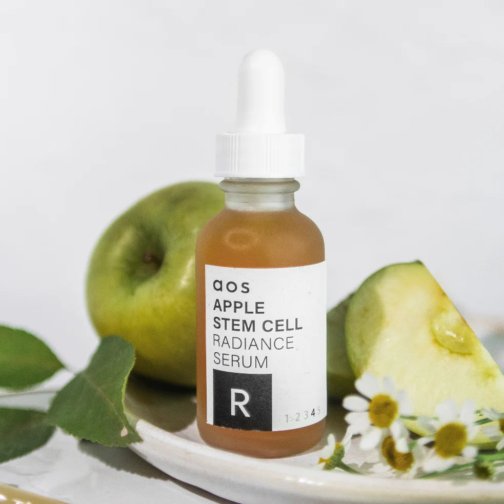 A bottle of apple stem cell radiance serum placed next to green apples and foliage on a ceramic plate.