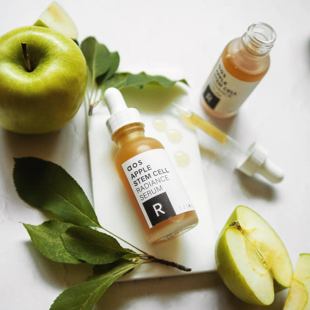 Bottles of apple stem cell serum surrounded by fresh apples and green leaves.