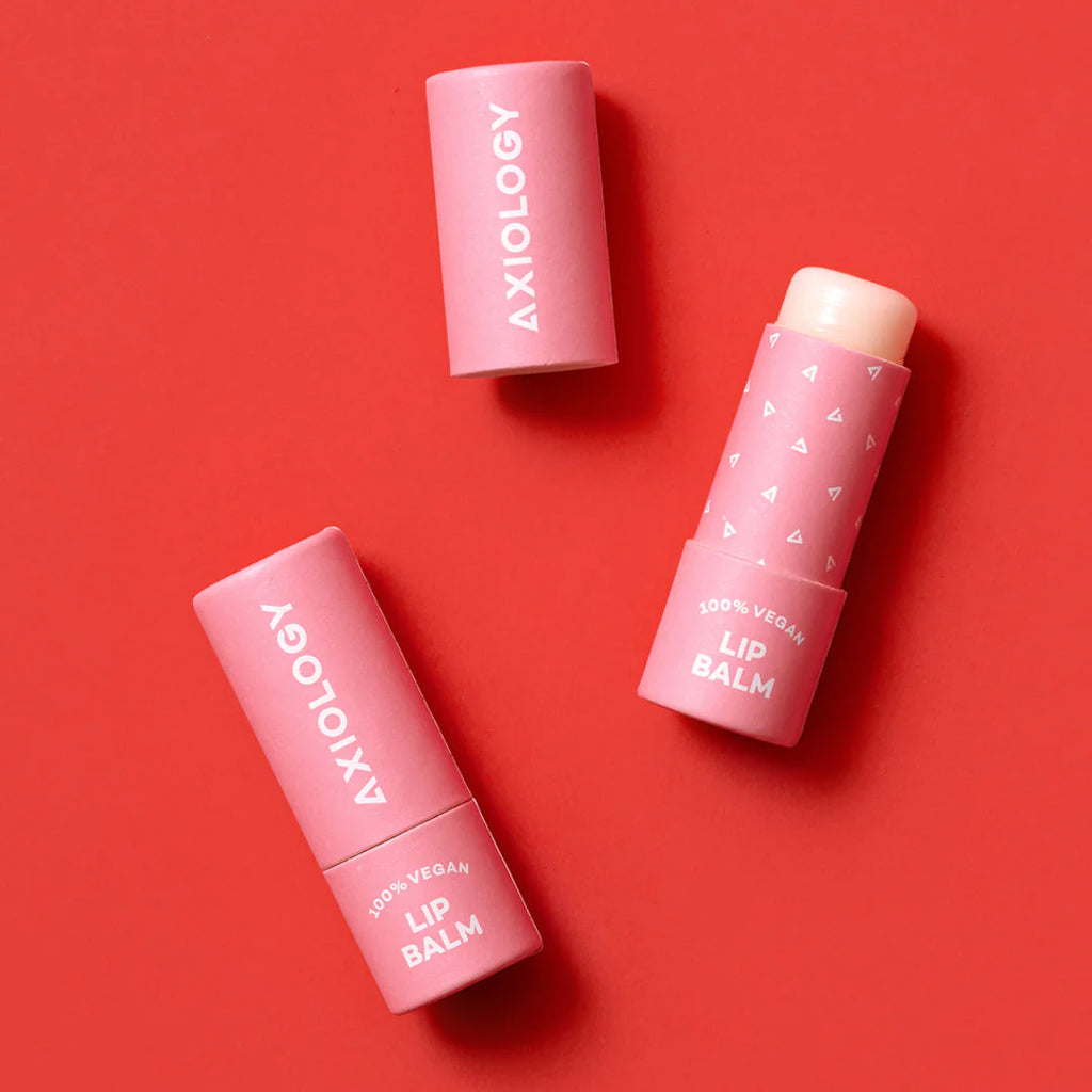 Three pink Axiology Vegan Lip Balm tubes labeled on a red background, with one tube opened revealing the product.