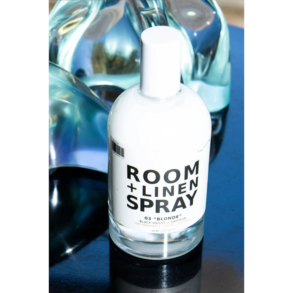 Bottle of room and linen spray with the scent labeled "03 blonde" displayed in front of blurred glass objects.