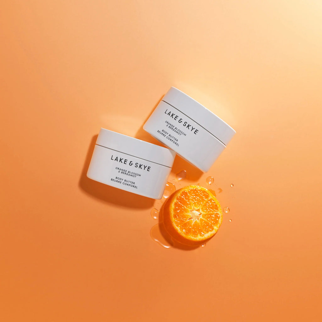 Two jars of lake & skye beauty products with a slice of orange and drops of liquid on an orange background.