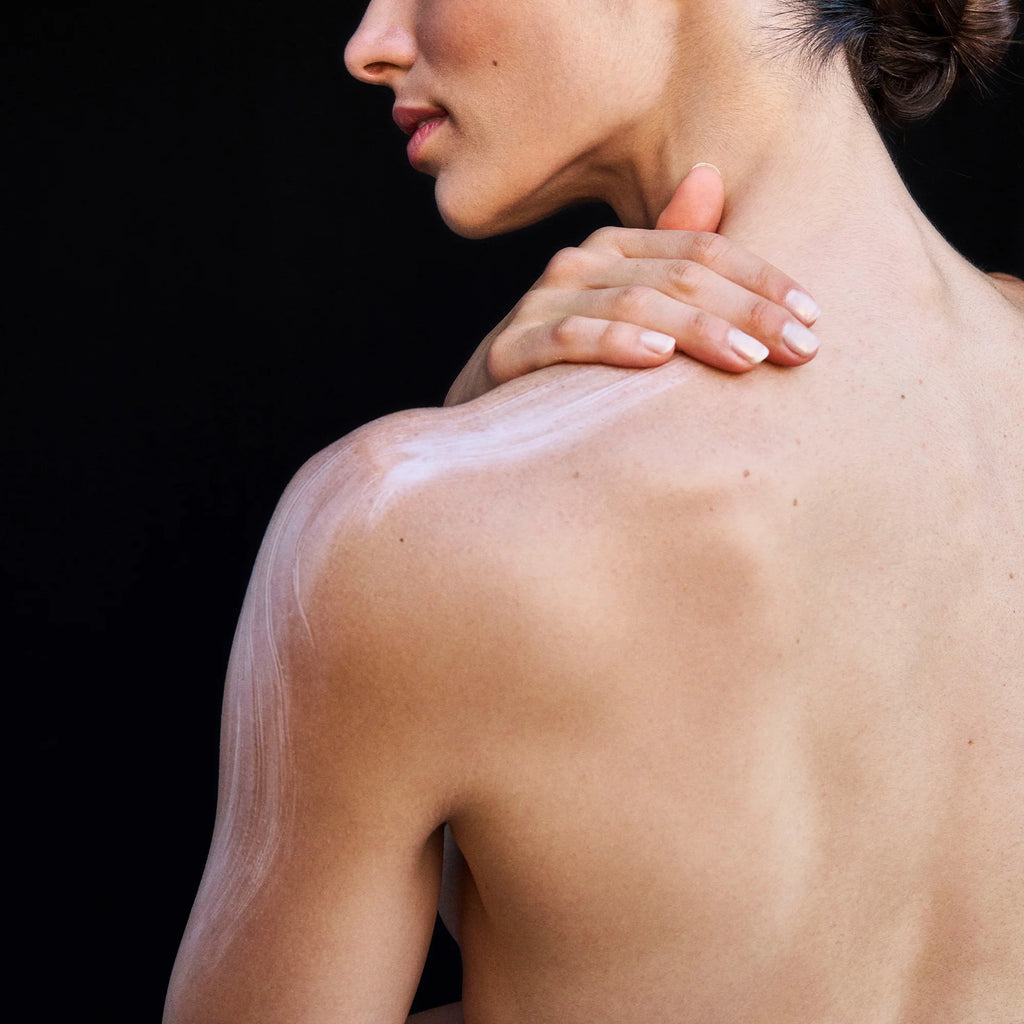 A person with bare shoulders applying lotion to their skin.
