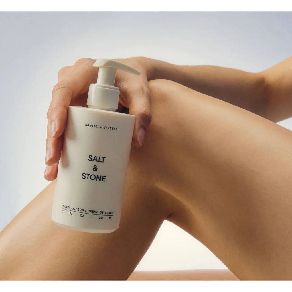 A person holding a bottle of salt & stone body lotion between their knees.