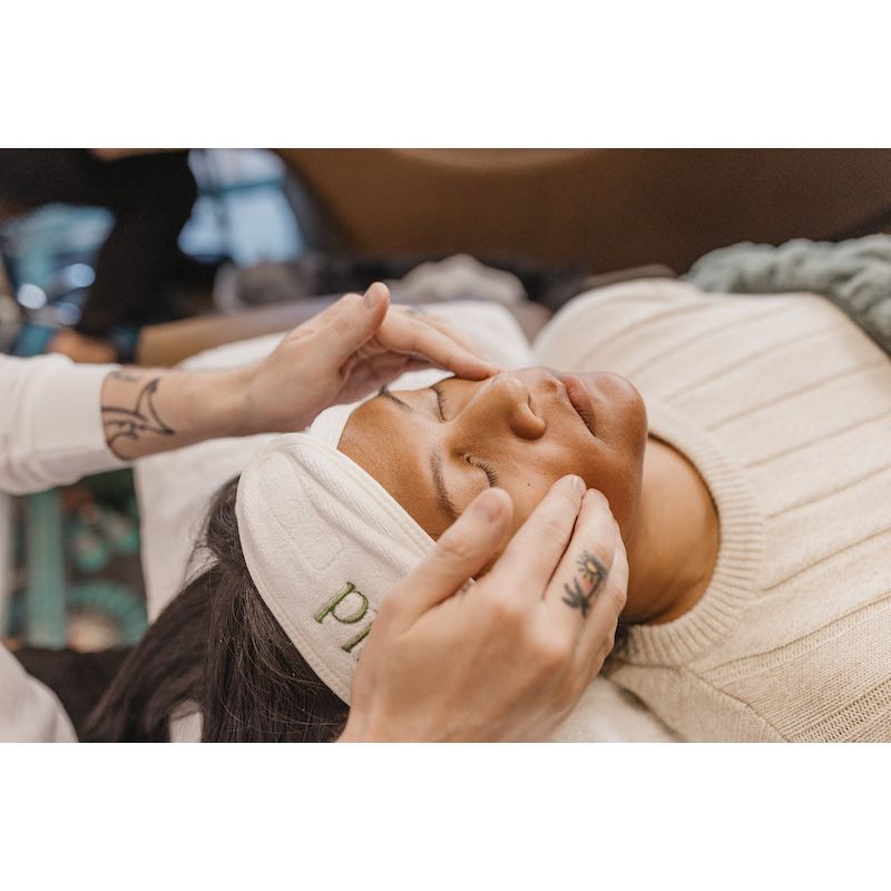 A person lies down with a headband on, enjoying a soothing Berlin Skin Matcha Mini Facial while another individual with visible tattoos massages their face using Berlin Skin products.