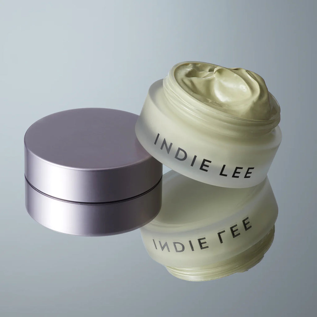 A jar of Indie Lee Color Balancer skincare cream with its lid off, showcasing the Tiger Grass color-correcting product inside, on a reflective surface.