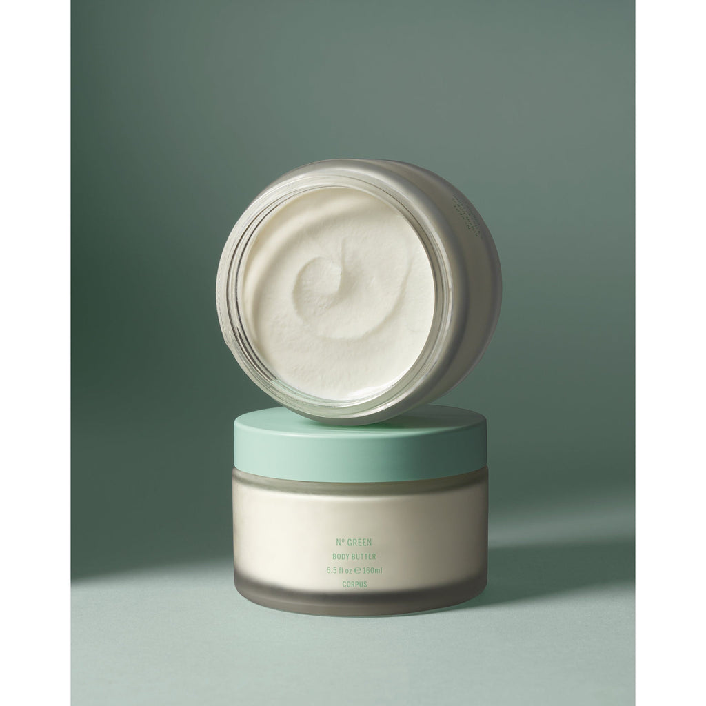 Two jars of Corpus body butter, one closed on a light green background the second sitting on top on it's side with the lid off exposing the white body butter inside