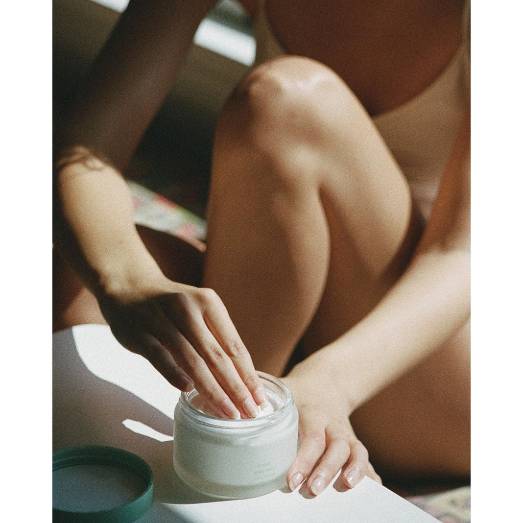 Female model sitting down with a white tank top on with her hand scooping corpus body butter out of the jar