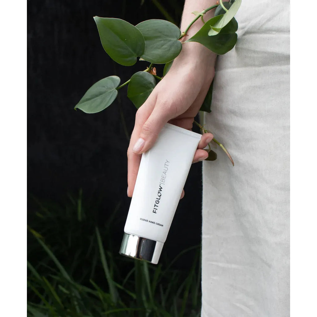 Person holding a tube of fenty beauty cream with green plant leaves protruding from the cap against a black and natural backdrop.