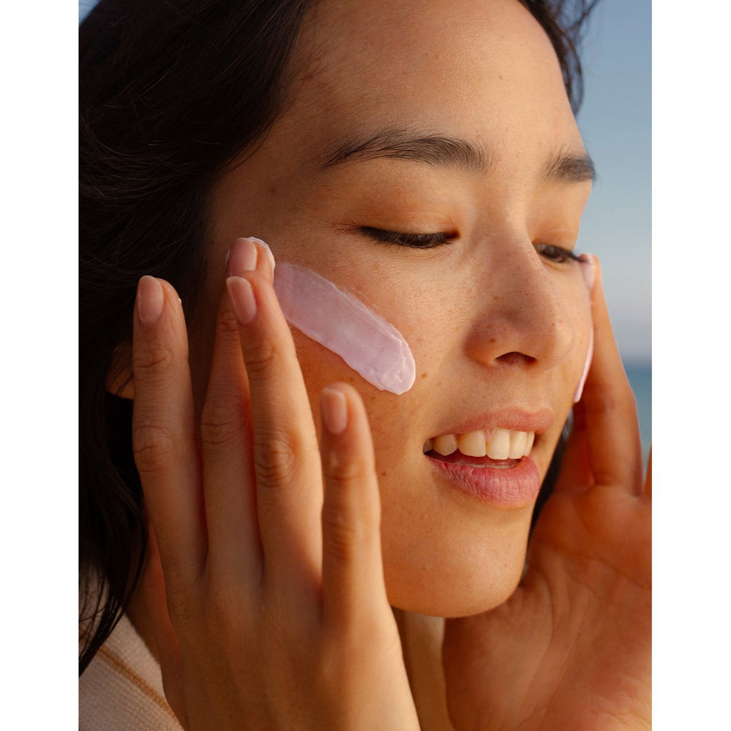 A woman gently holds a pink gua sha tool to her cheek as part of a skincare routine.