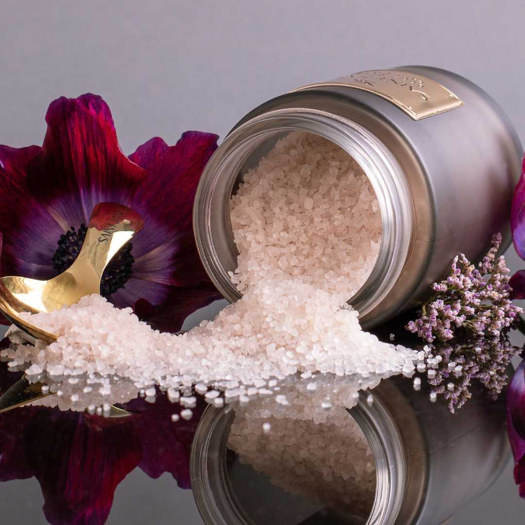 A jar of bath salts spilled over a reflective surface, surrounded by dark red petals and purple flowers.