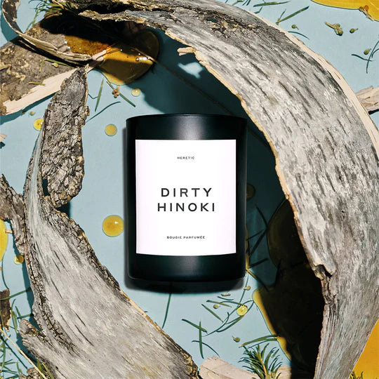 A scented candle labeled "dirty hinoki" displayed amidst rustic wooden branches on a patterned background.