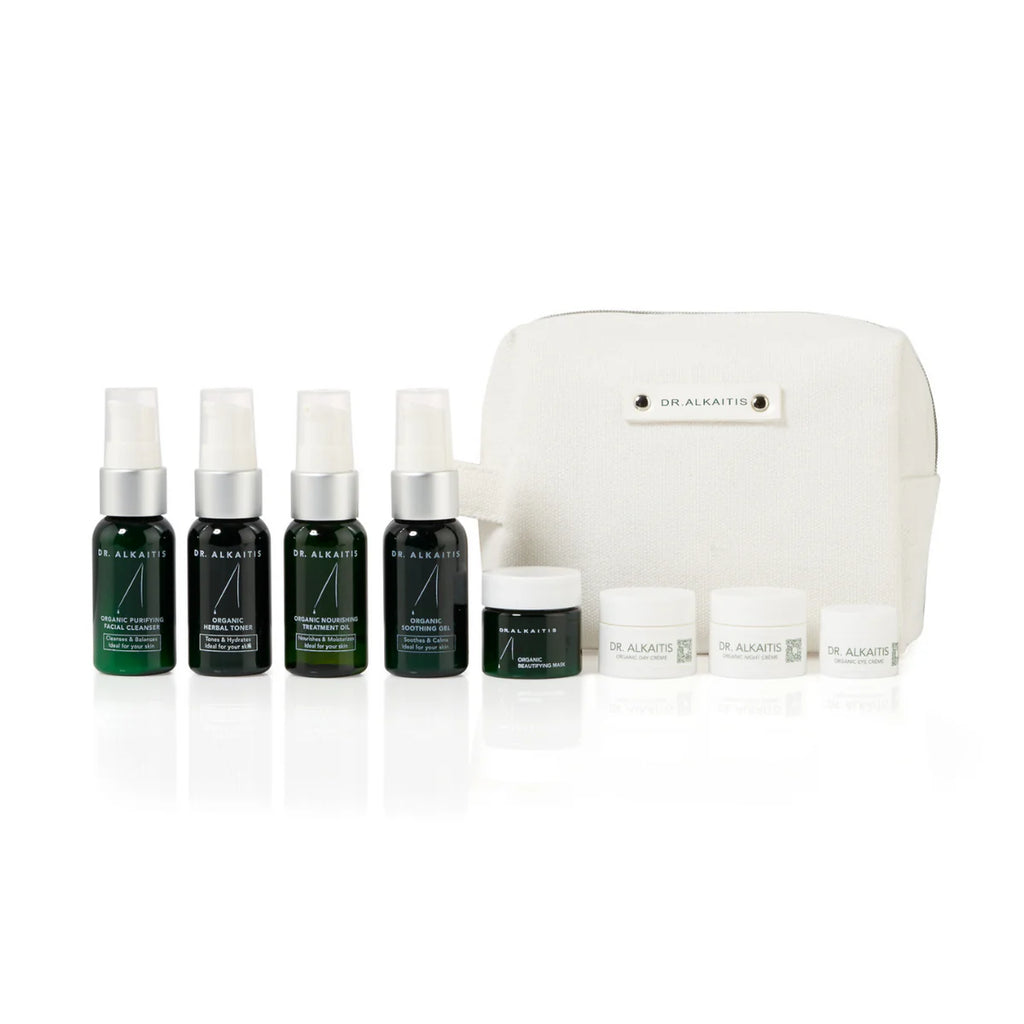 A TSA-approved, Dr. Alkaitis Organic Travel Kit Deluxe comprising skincare products in bottles and jars, beautifully arranged in front of a white cosmetic travel bag. Ideal for eco-conscious travelers.