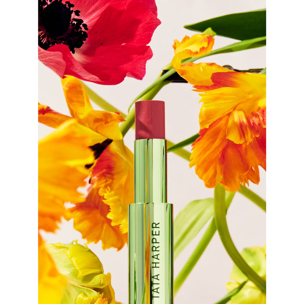 A tube of tata harper lip balm set against a backdrop of vibrant red and yellow flowers.