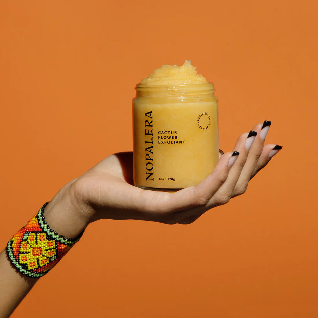 A hand with black nail polish holding a jar of nopalera cactus flower exfoliant against an orange background.