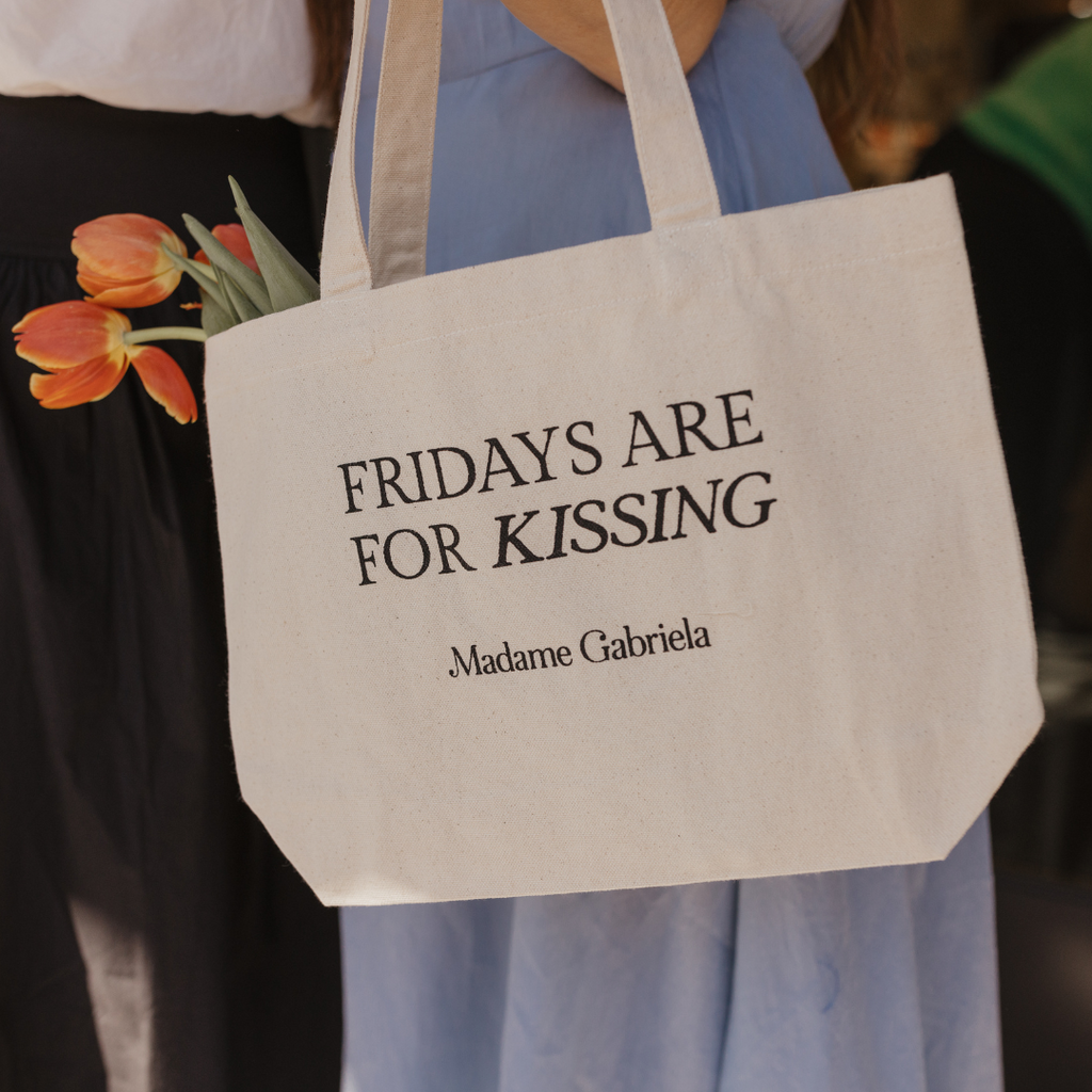 A person is holding an organic bag with the text "FRIDAYS ARE FOR KISSING" and "Madame Gabriela" printed on it. Orange tulips are partially tucked into the Fridays Are For Kissing Madame Gabriela Tote, making it perfect for everyday errands.