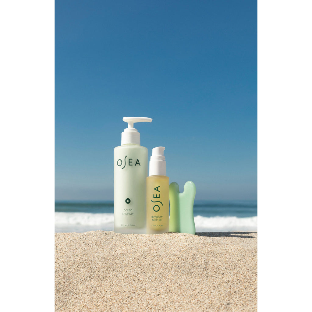 Two skincare products positioned on sandy beach with ocean waves in the background.