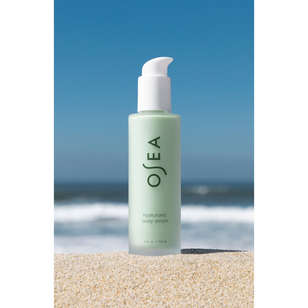 A bottle of Osea Hyaluronic Body Serum on a sandy beach with ocean waves in the background.