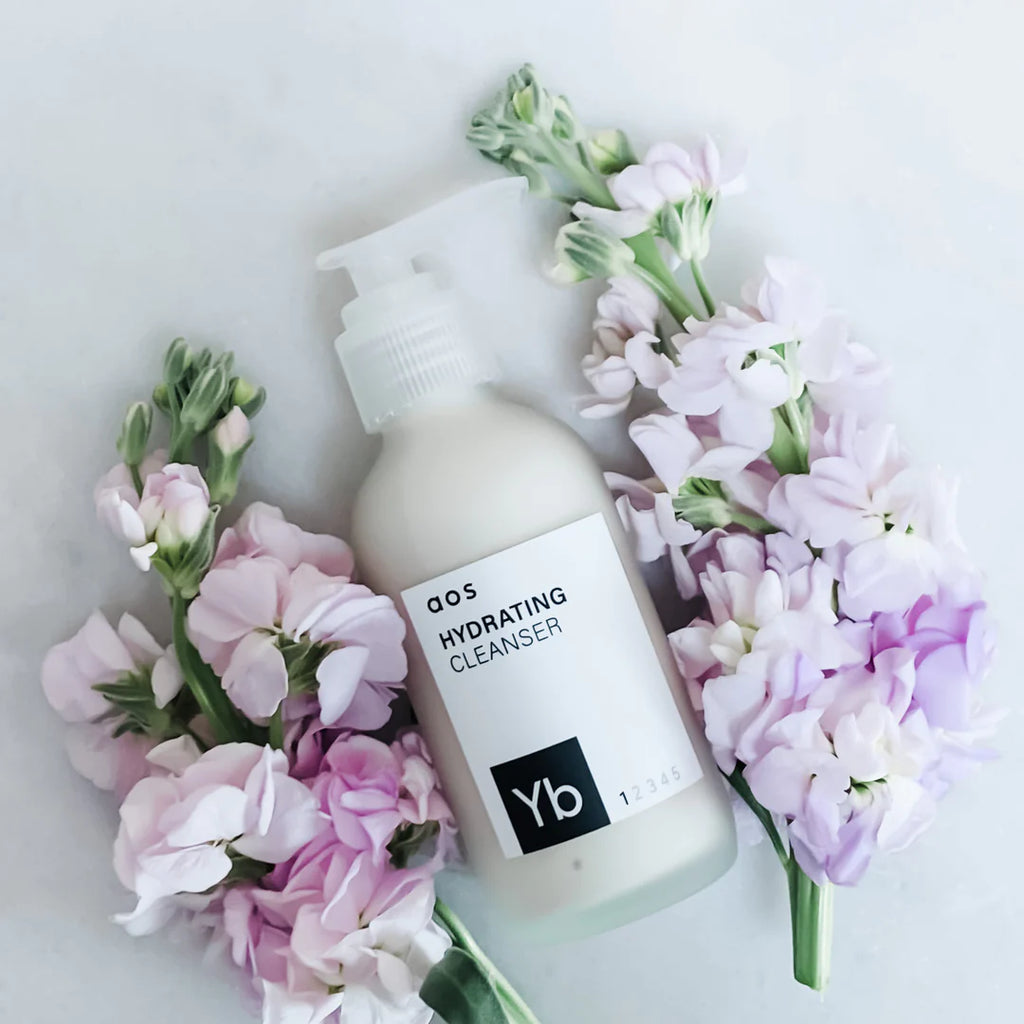 A bottle of hydrating cleanser surrounded by delicate pink flowers on a light background.