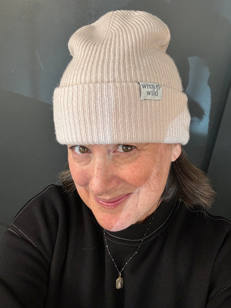 A person wearing a cream-colored Wren and Wild Cashmere Beanie smiles at the camera. They have a black top and a necklace.