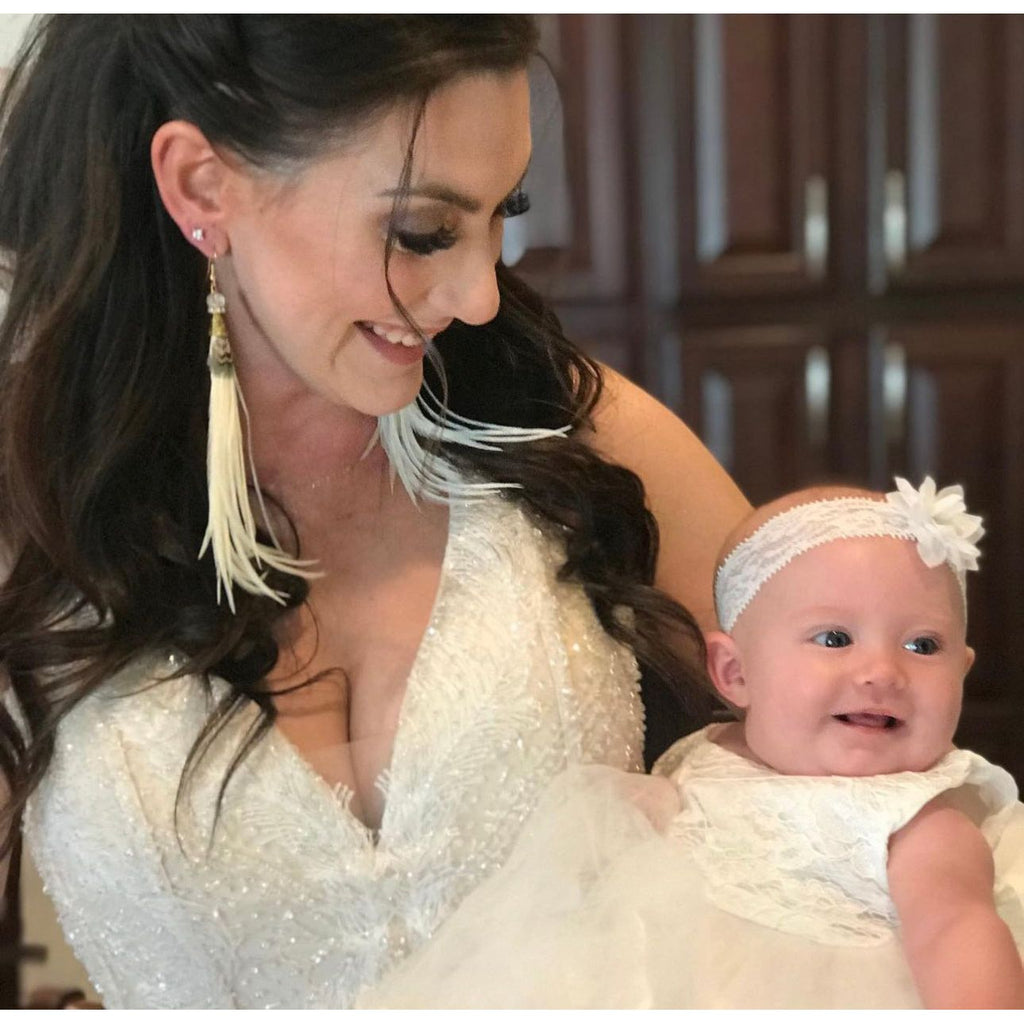 A woman in a white dress with tassel earrings smiles down at a baby in a white outfit and headband.