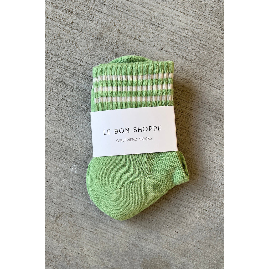 A pair of green and white striped socks with a label reading "le bon shoppe girlfriend socks" on a gray textured surface.