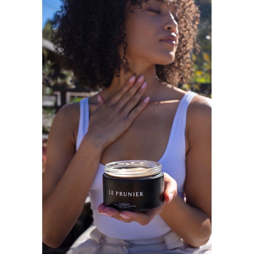 A person holding a jar of "le prunier" beauty product while touching their neck with sunlight illuminating their profile.