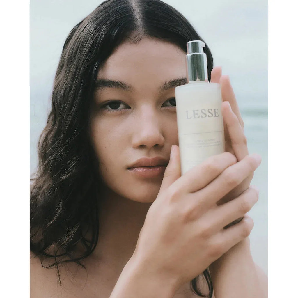 A woman holding a bottle of LESSE Calming Cleanser close to her face, with a blurred ocean background. Perfect for those with reactive skin.