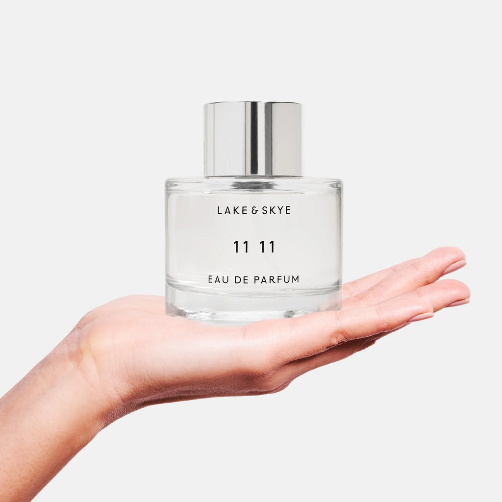 A hand presenting a clear bottle of lake & skye 11 11 eau de parfum against a white background.