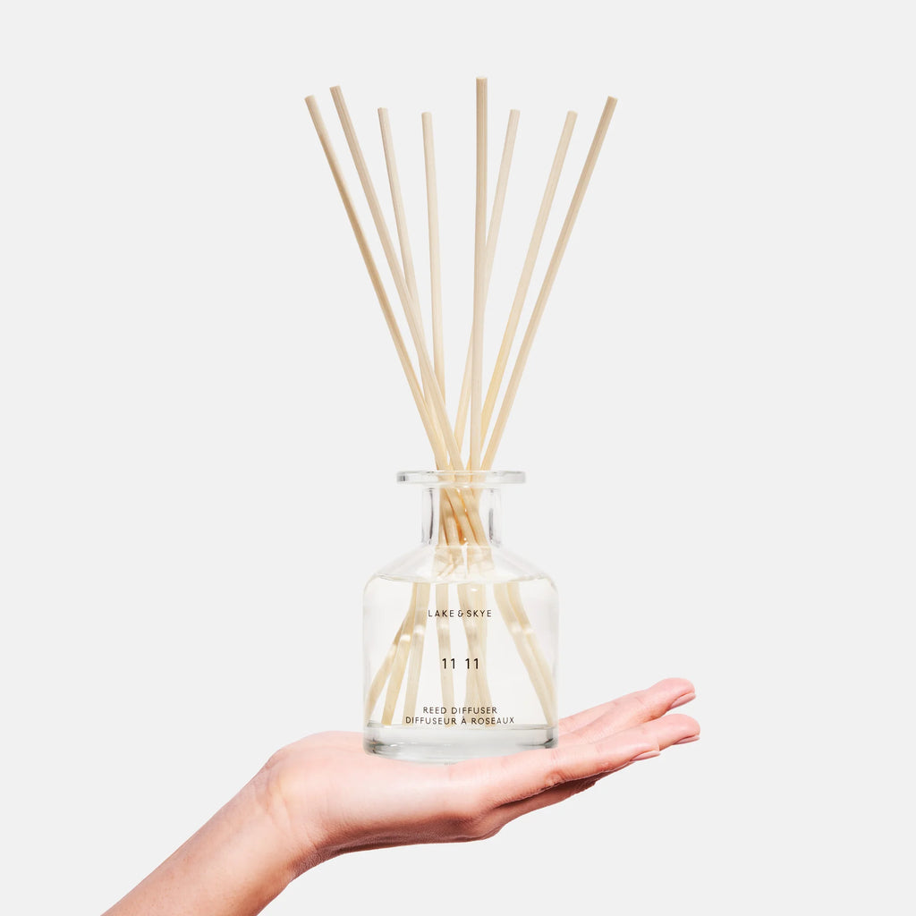 A hand holding a clear diffuser bottle with reed sticks.