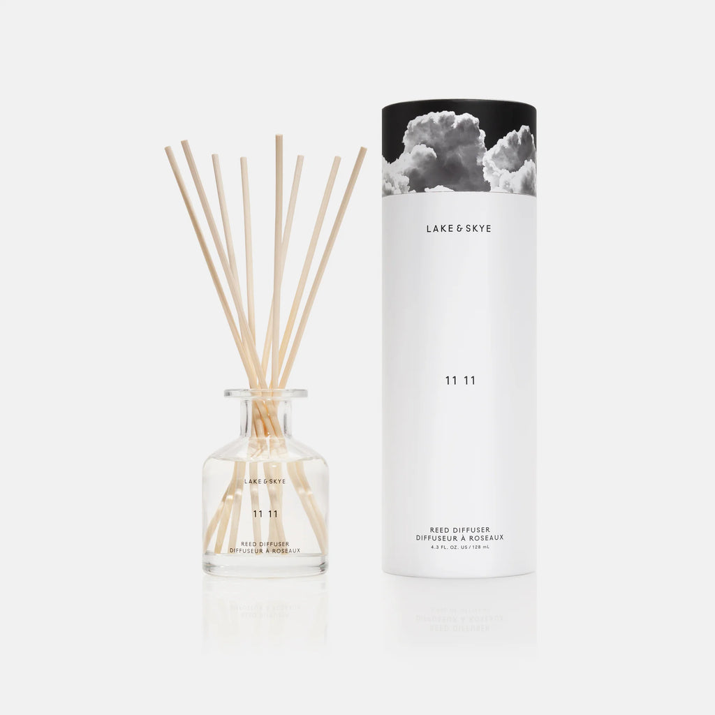 A reed diffuser with wooden sticks inserted into a clear bottle, alongside its cylindrical packaging with "lake & skye 11 11" branding.