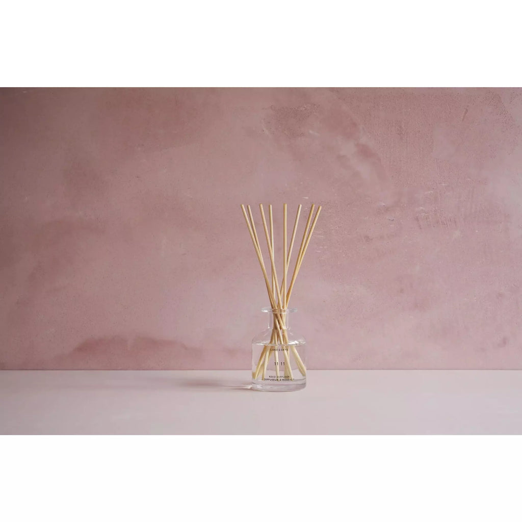 A reed diffuser with several sticks in a clear glass jar on a pink surface against a pink textured backdrop.