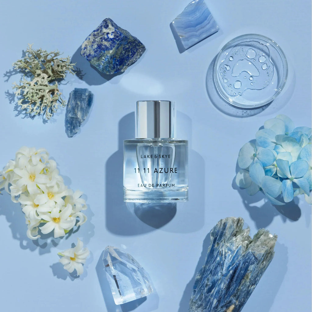 Flat lay composition with a bottle of lake & skye 11 11 azure perfume surrounded by blue crystals, flowers, and a container of cream on a pale blue background.