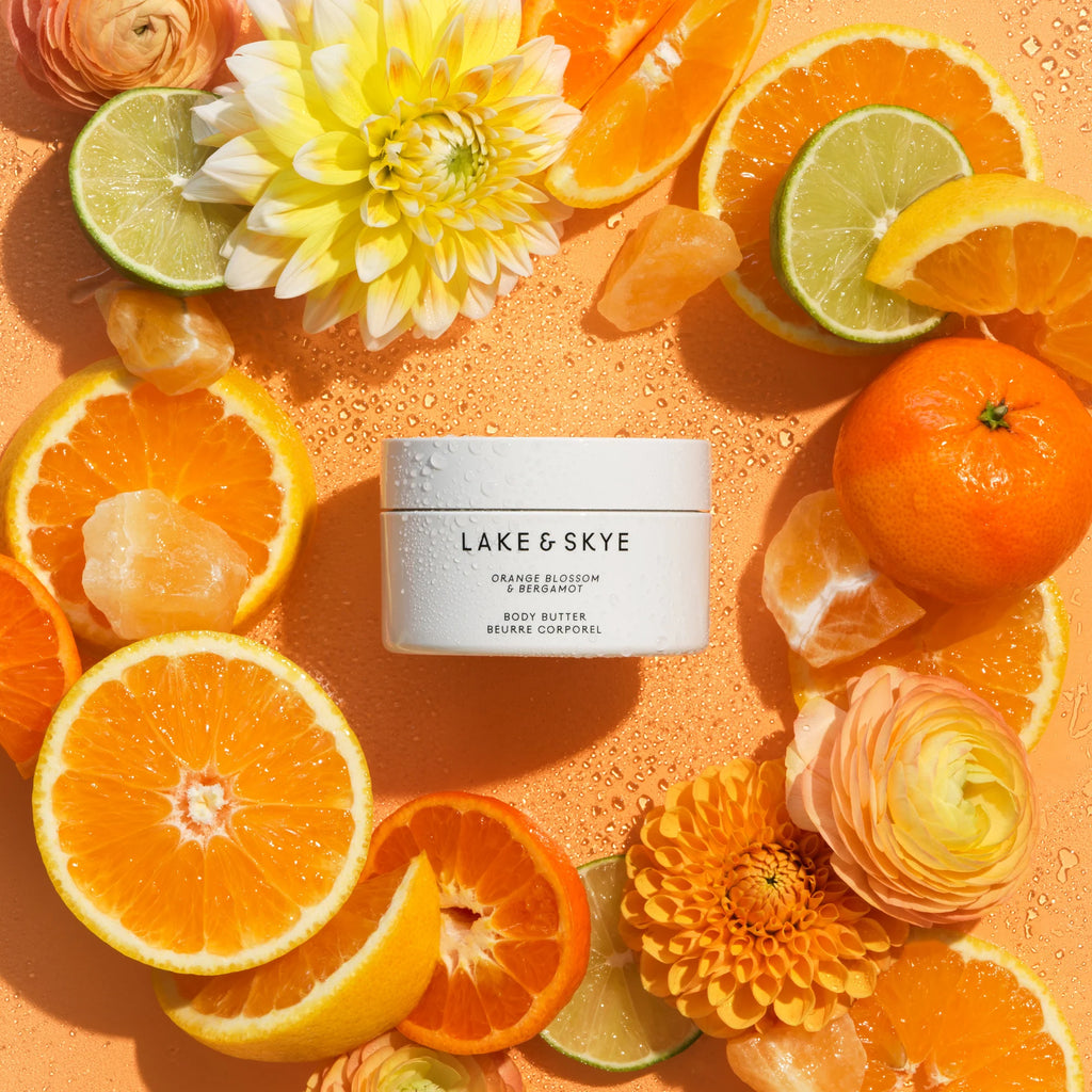 A jar of lake & skye orange blossom body butter surrounded by fresh citrus slices and flowers on an orange backdrop.