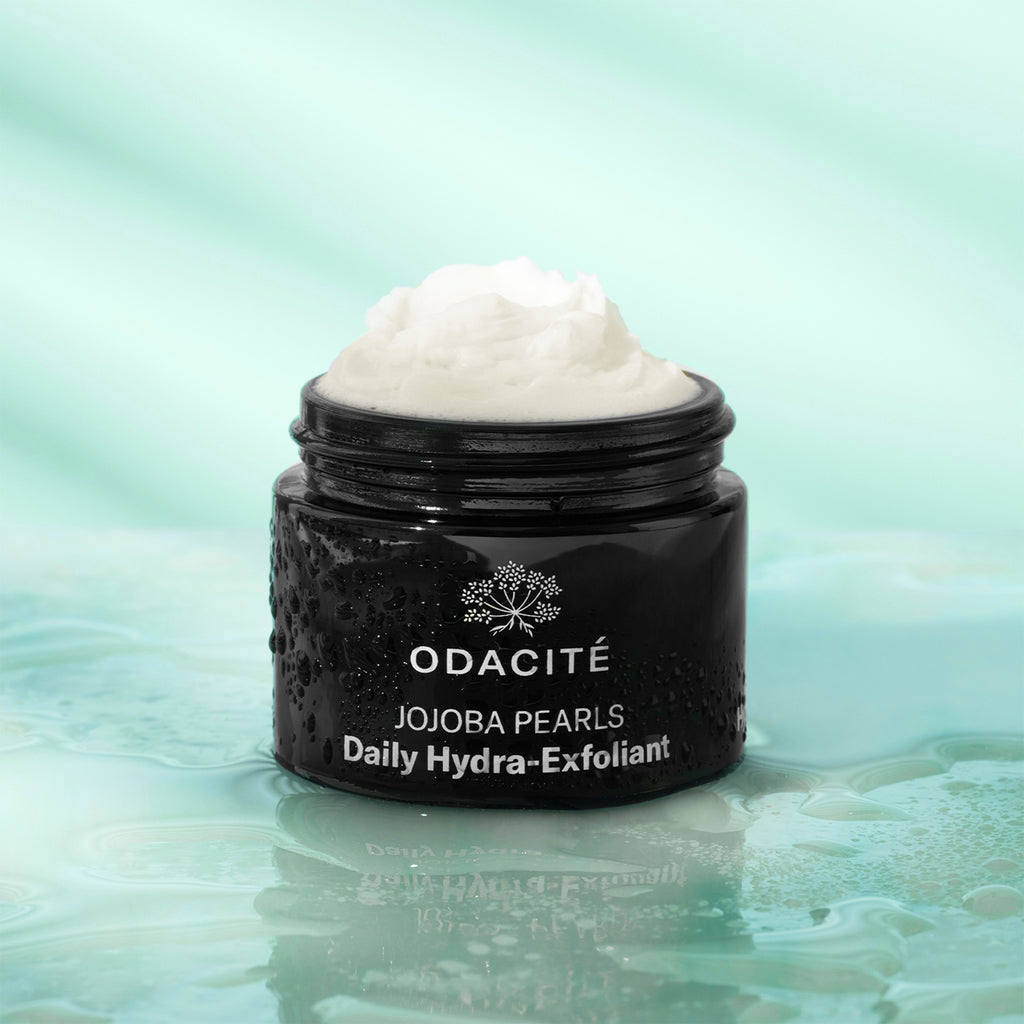 A jar of odacite jojoba pearls daily hydra-exfoliant with a dollop of cream on top, displayed against a watery background.