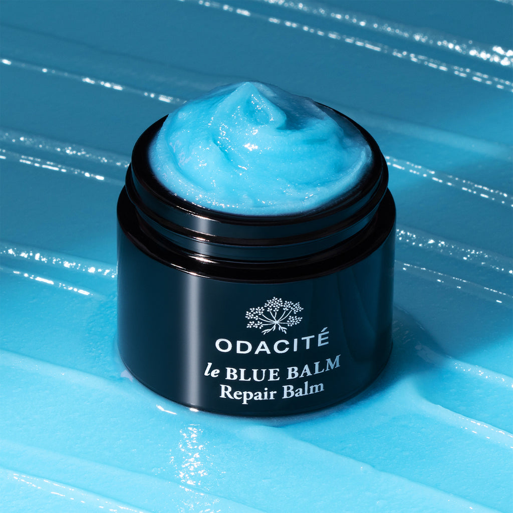 Jar of odacite le blue balm on a blue textured surface with the lid off, showing the blue product inside.