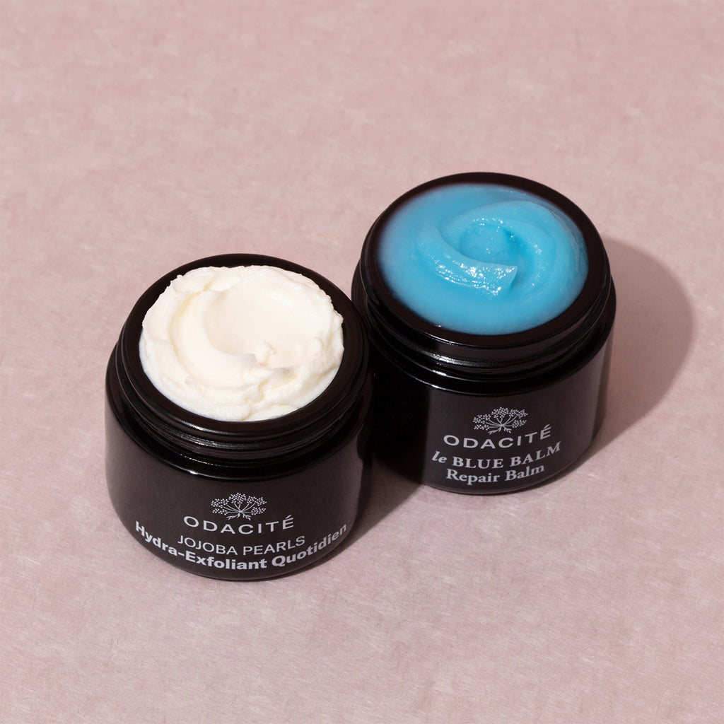 Two jars of odacite skincare products open to show their contents; one with a creamy white texture and the other with a blue balm.