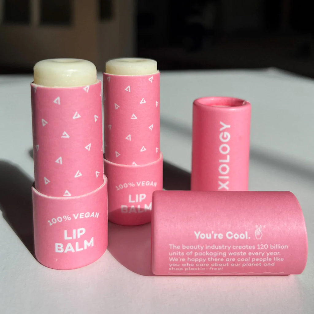 Three pink Axiology Vegan Lip Balms with caps are displayed in natural lighting; one is laying sideways with a supportive environmental message visible. These handmade in USA, cruelty-free lip care products are perfect for eco-conscious consumers.