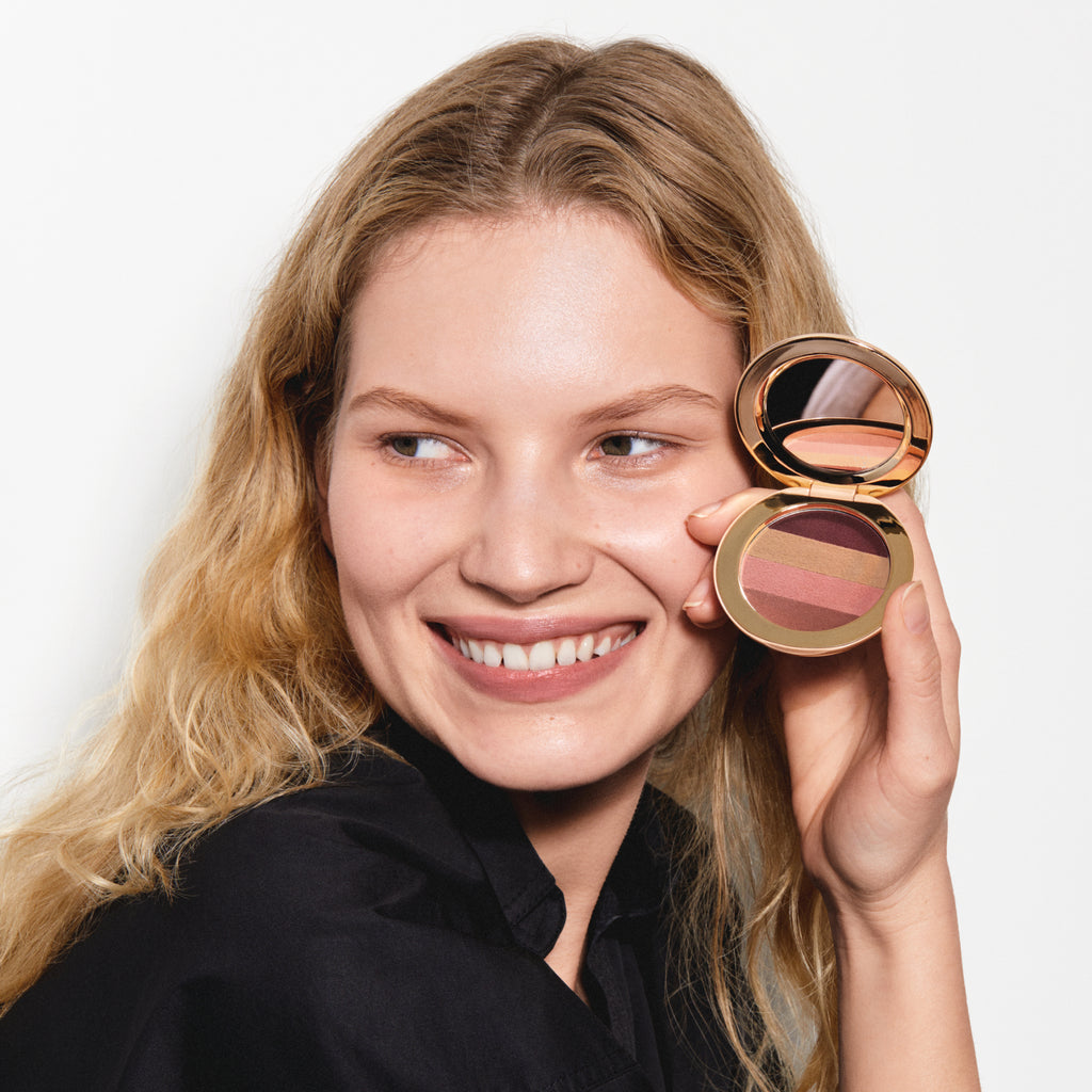 Woman smiling while holding a compact makeup palette near her face.