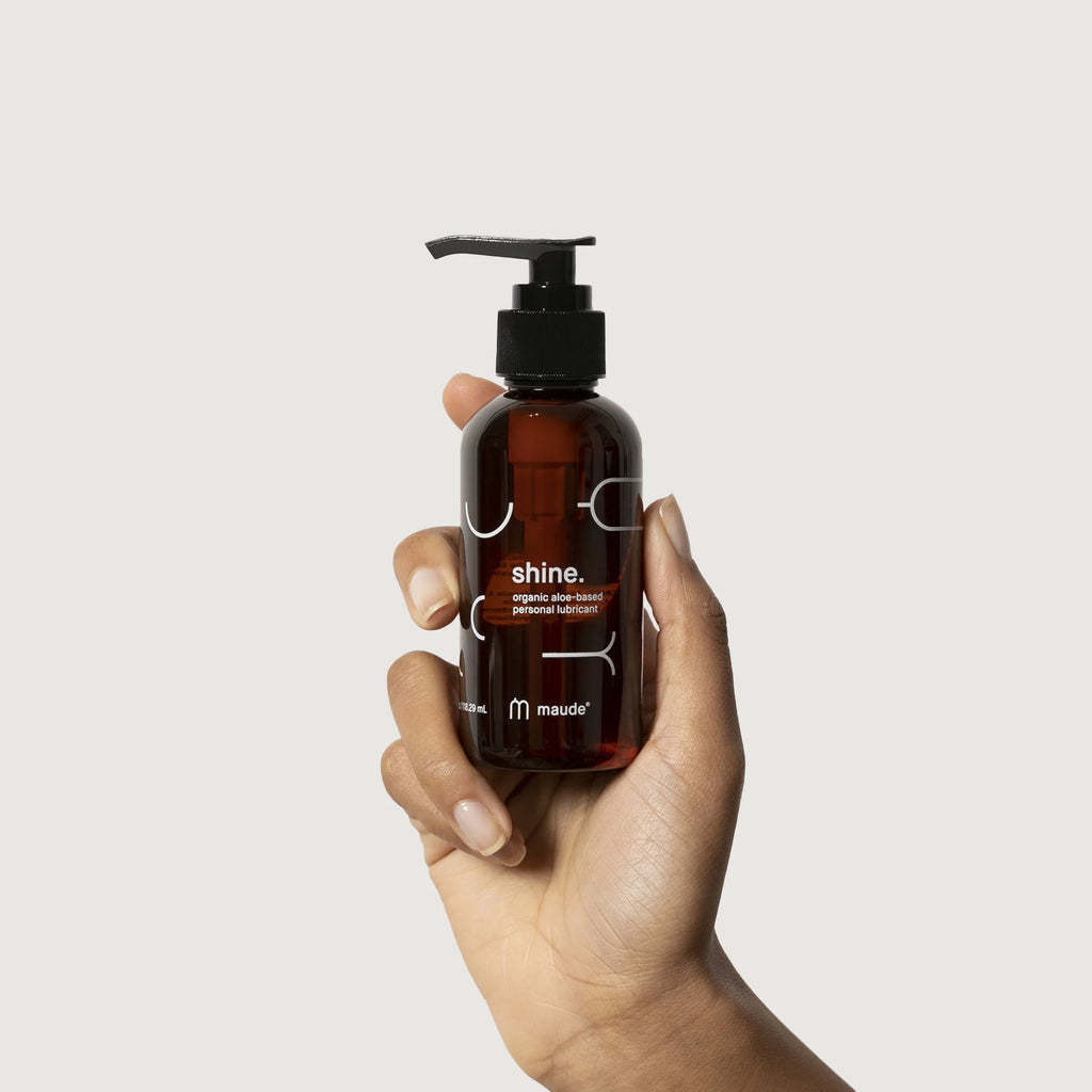 A hand holding a bottle of 'shine' organic lubricant by maude against a plain background.