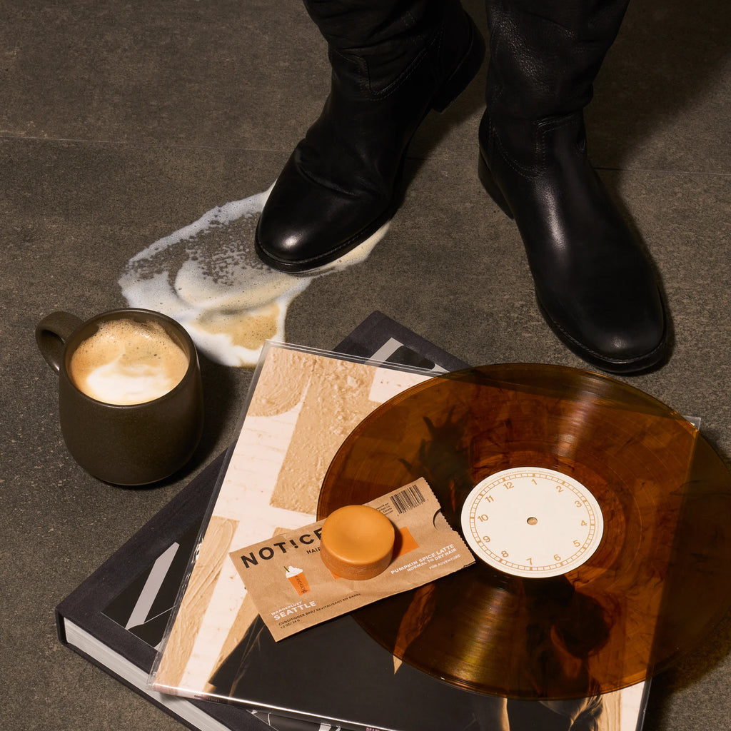 A spilled pumpkin spice latte rests beside a vinyl record, a NOTICE HAIR CO. SEATTLE Conditioner Bar in its packaging, and a pair of black boots on the gray floor.