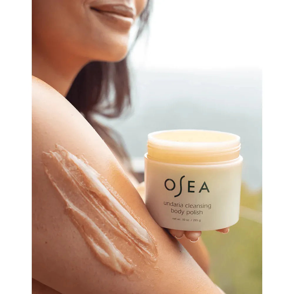 A person applying osea undaria cleansing body polish on their arm, smiling in the background.