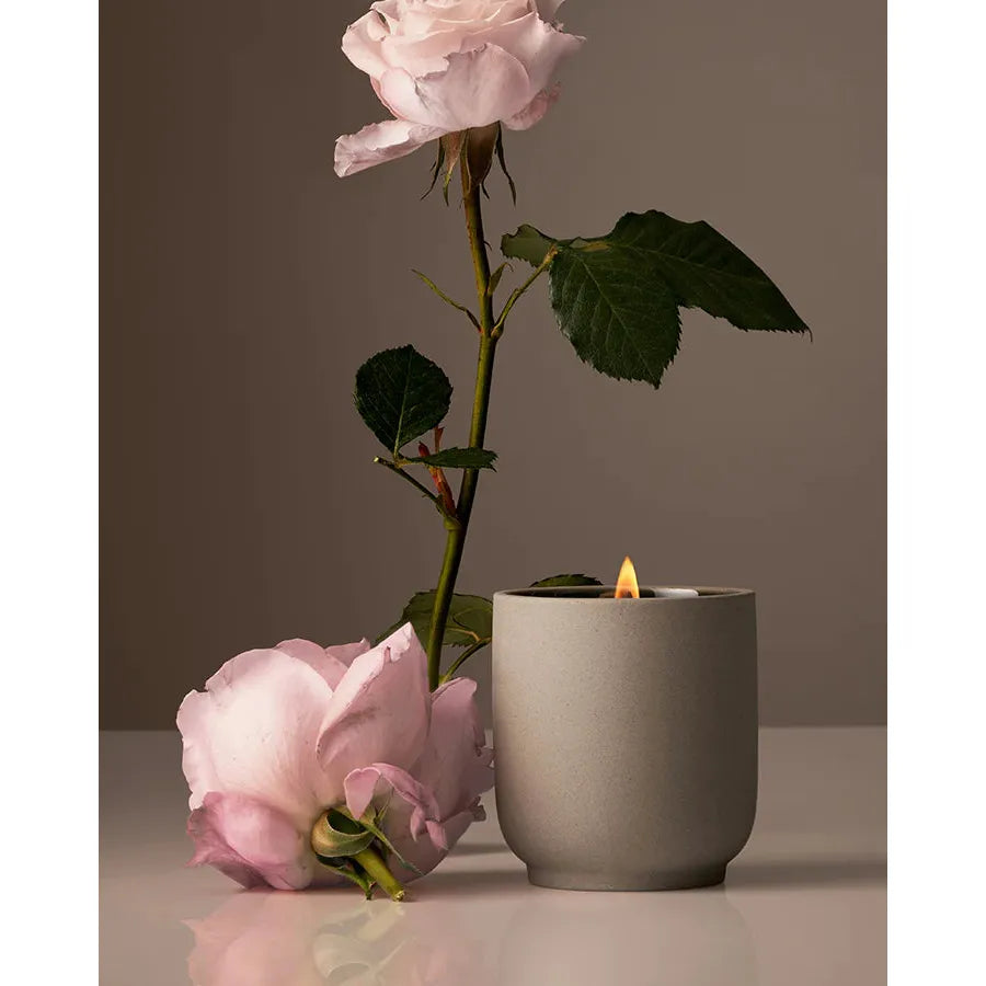 A lit candle in a gray holder beside two pink roses, one standing in a vase and the other lying on the surface.