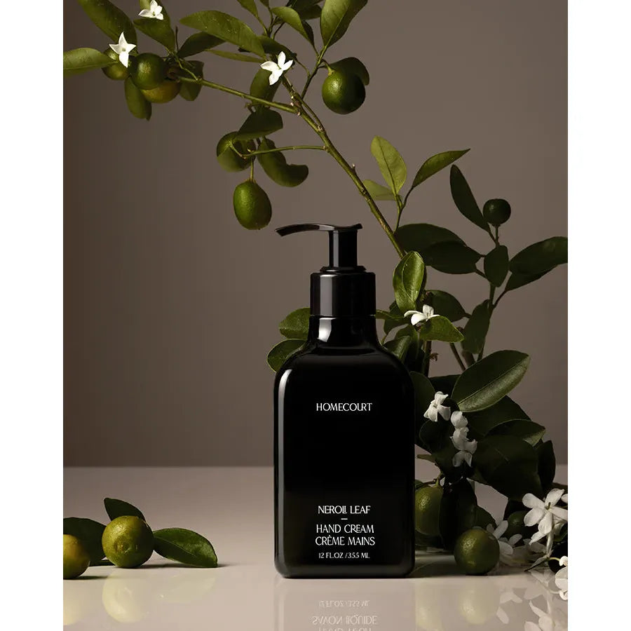 A black dispenser bottle of Homecourt Hand Cream on a beige background, surrounded by dark green leaves and small white flowers.