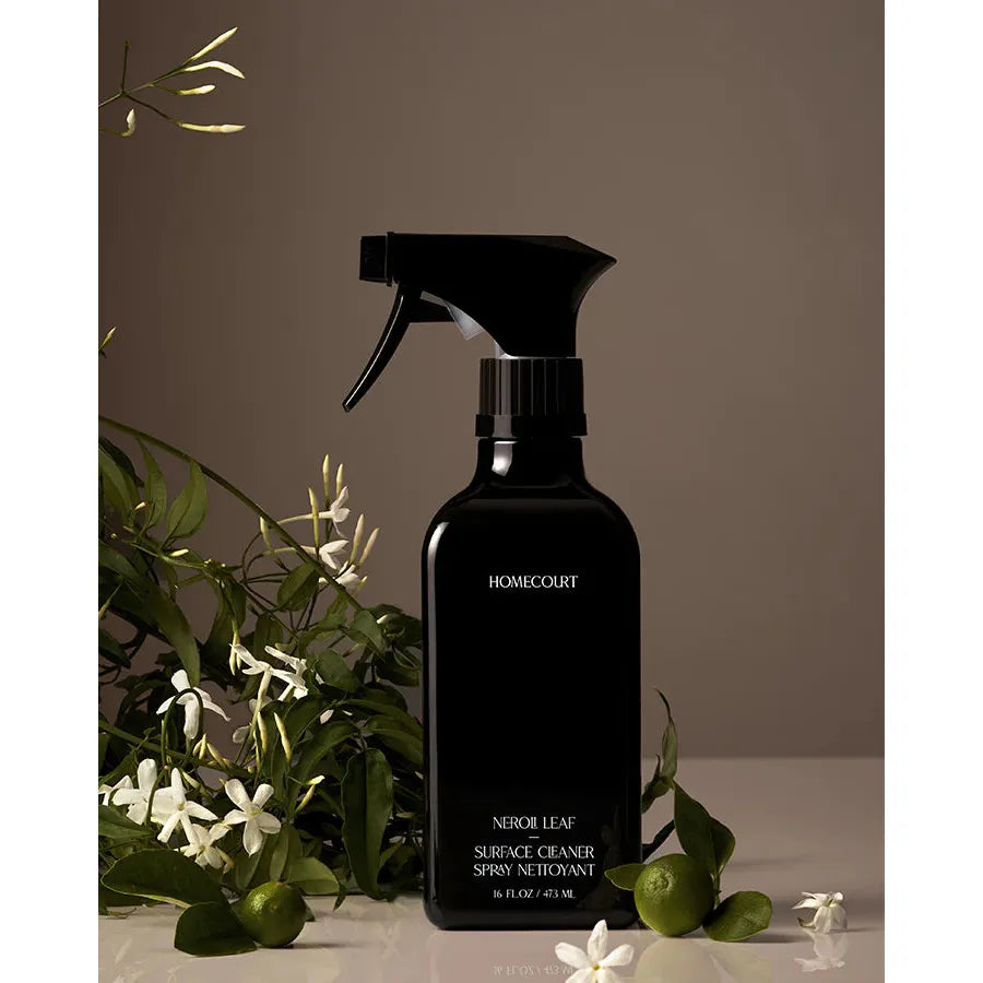 Black spray bottle labeled "Homecourt Surface Cleaner" with a coconut-derived degreaser on a brown background with scattered white flowers and green leaves.