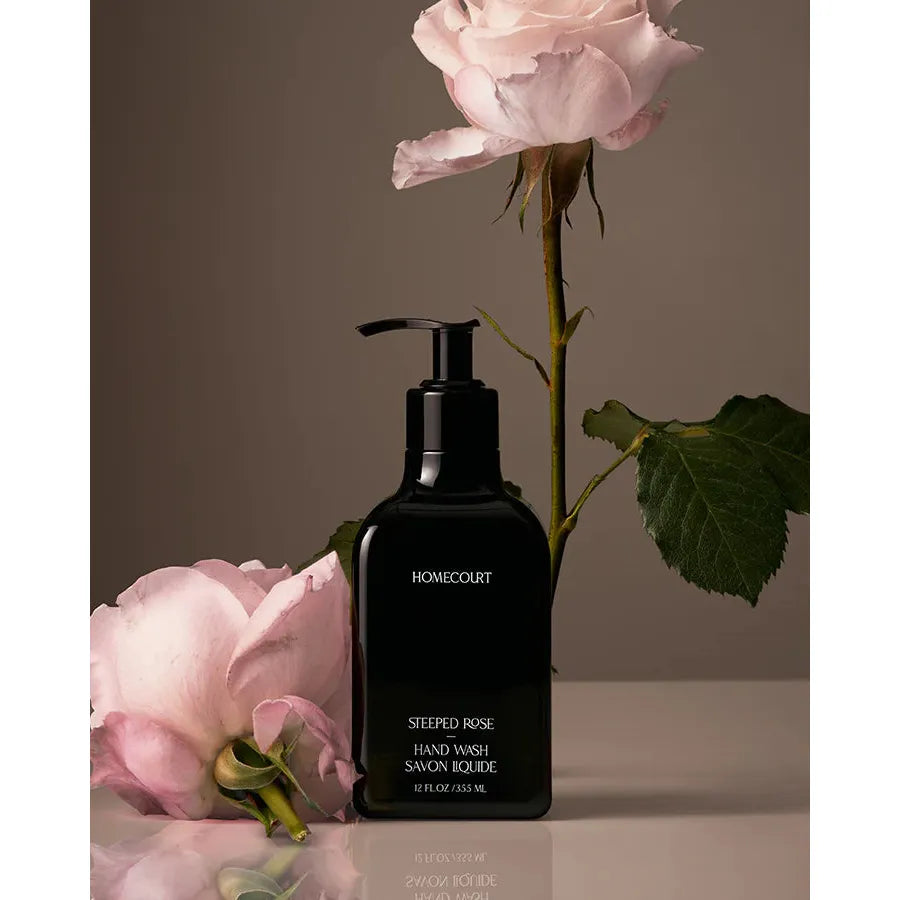 A bottle of homecourt's steeped rose hand wash on a neutral background with a rose stem leaning against it and a loose petal lying in front.